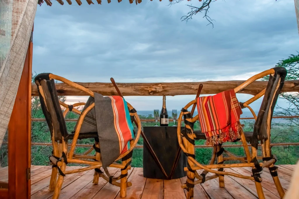 Two rustic wooden safari chairs draped with Maasai blankets facing champagne and glasses on a private balcony deck at Amani Eserian Ngorongoro, overlooking the crater landscape