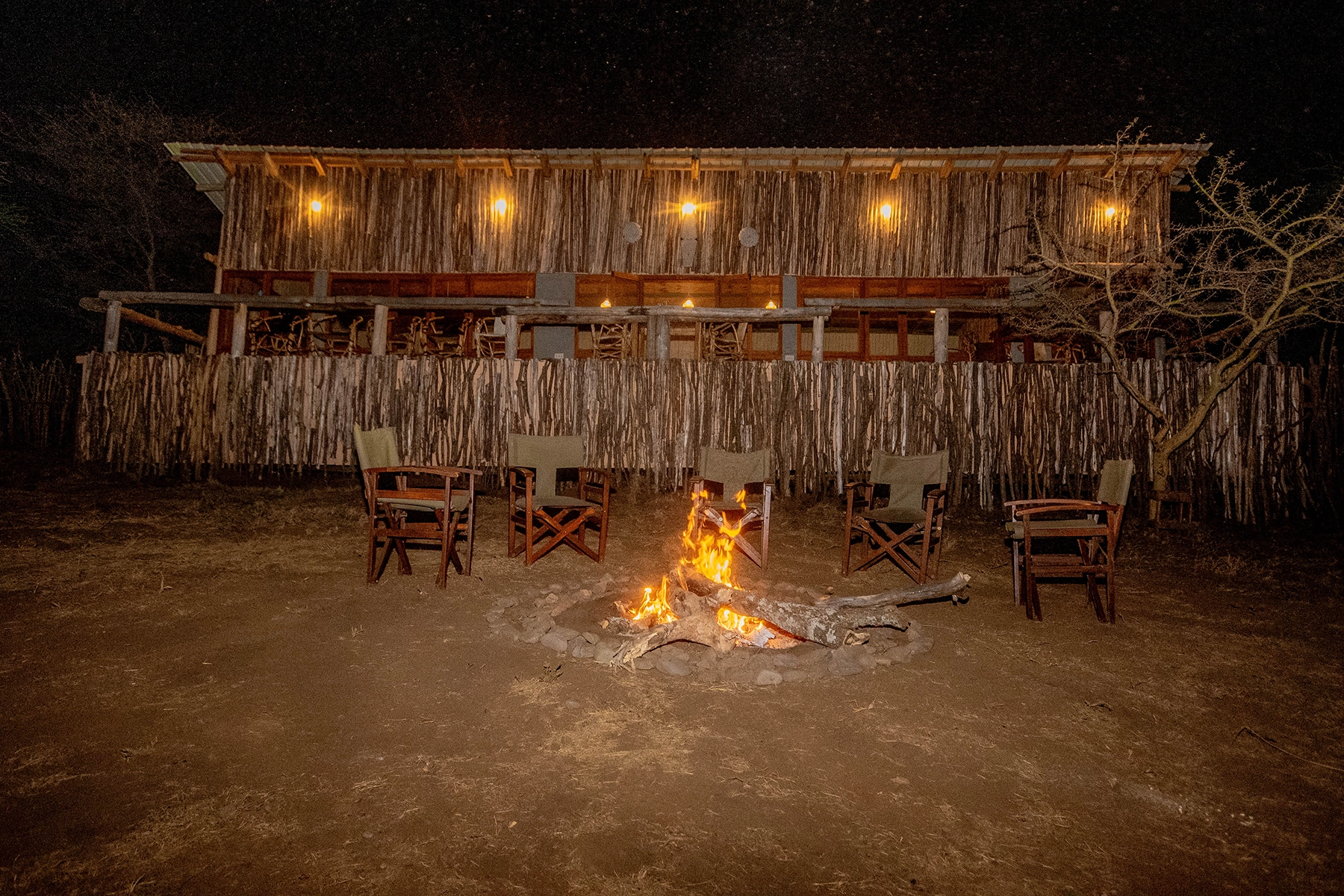 Evening campfire with safari director chairs arranged around an open fire in front of the rustic timber lodge building at Amani Eserian Ngorongoro under a starry night sky