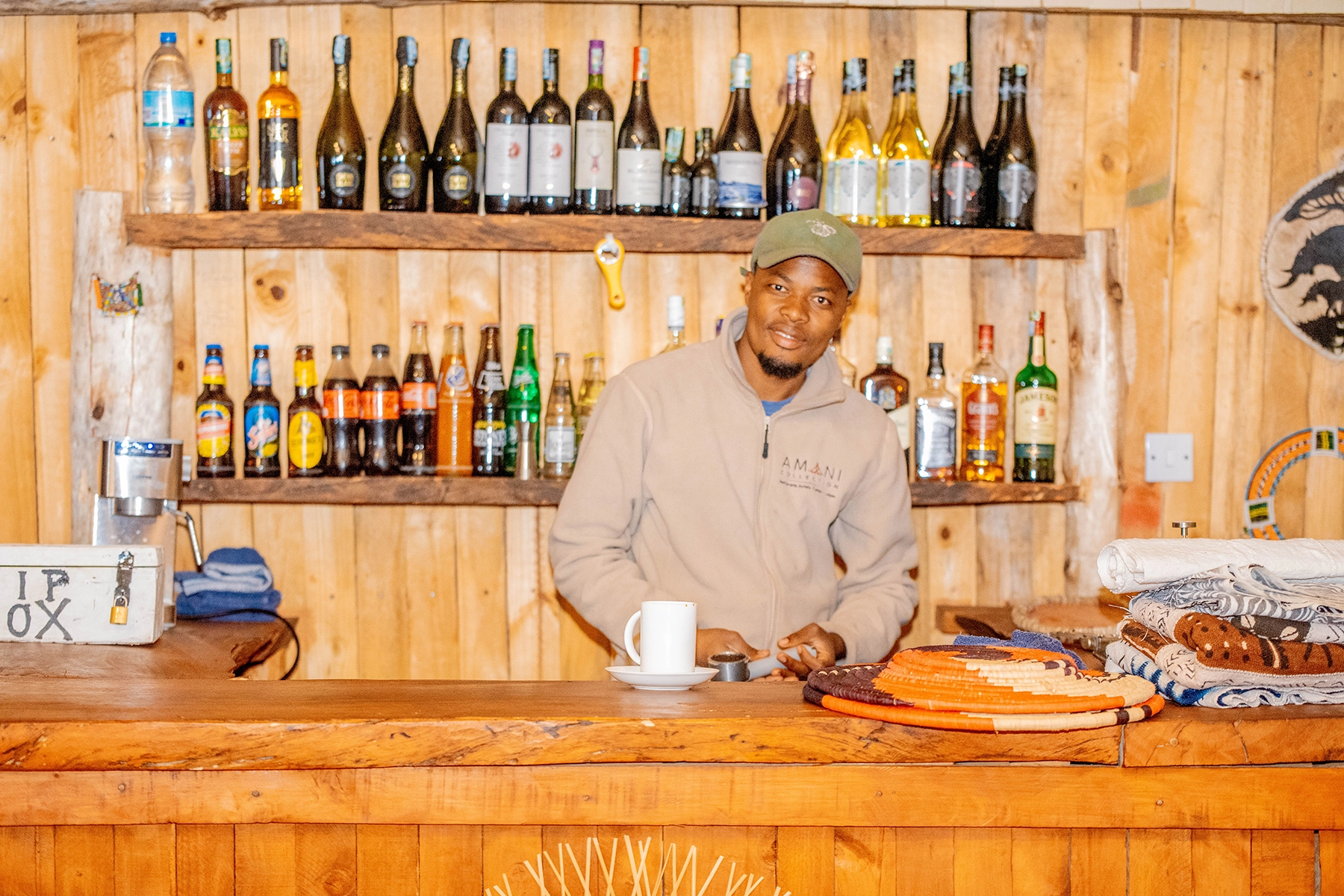 Friendly Amani Collection staff member behind the rustic wooden bar at Amani Eserian Ngorongoro, surrounded by a well-stocked selection of wines and spirits