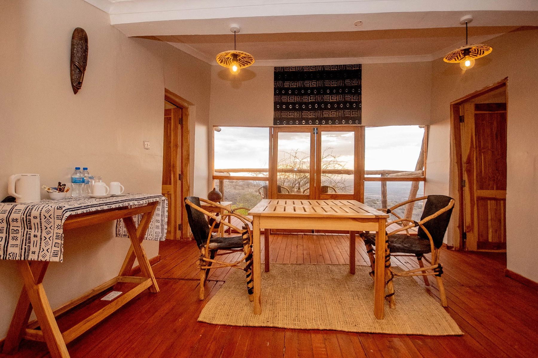 Safari suite lounge at Amani Eserian Ngorongoro with rattan chairs, wooden dining table, African mudcloth wall hanging, and panoramic window overlooking the Ngorongoro Crater