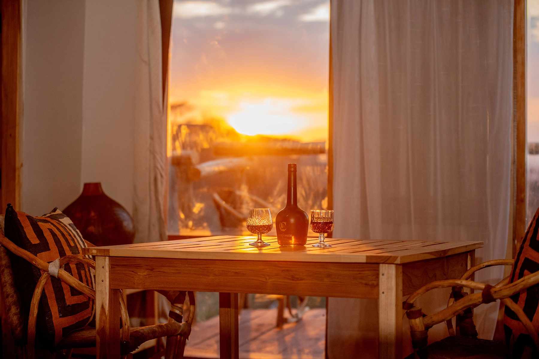 Dramatic golden sunset over the Ngorongoro Crater viewed through a lodge window at Amani Eserian, with drinks on a wooden table in the foreground