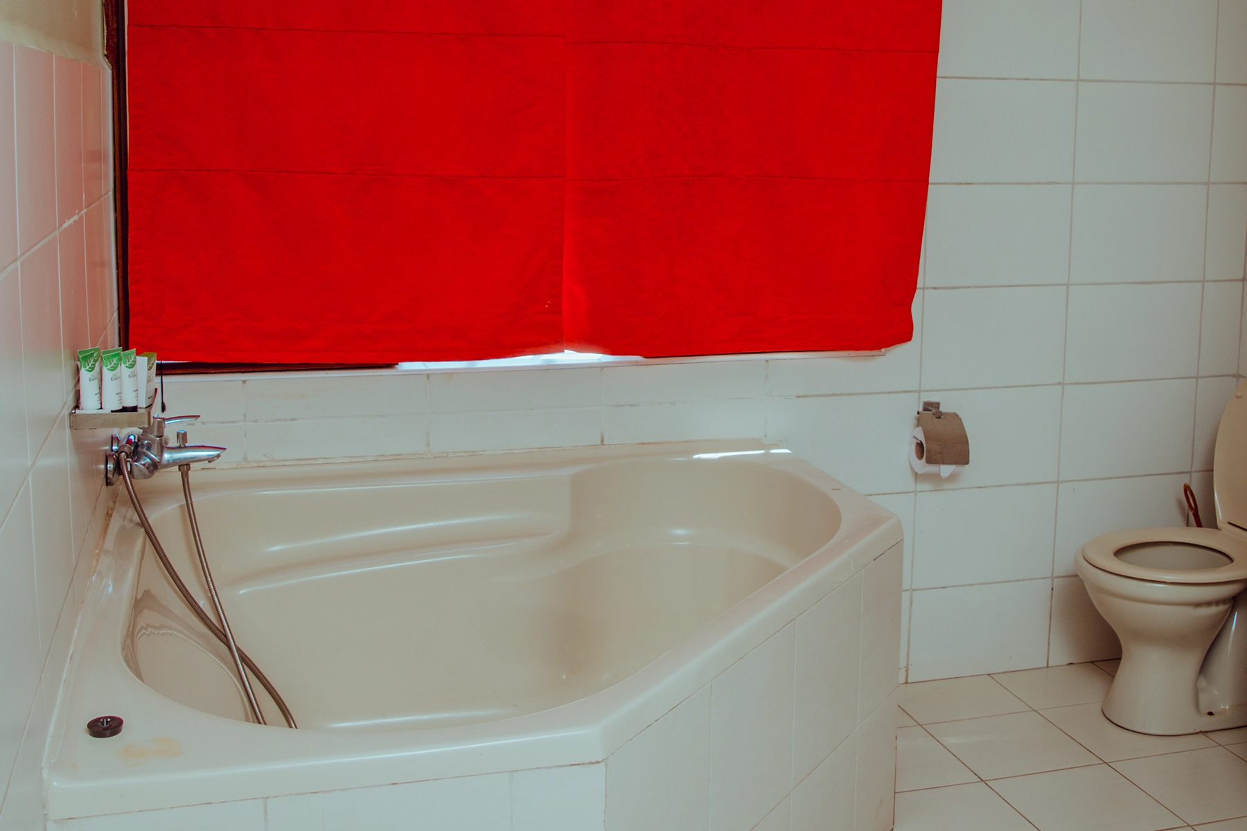 En-suite bathroom at Amani Farmhouse Tanzania featuring a corner soaking bathtub with chrome fittings, white tiled walls, and a red window blind