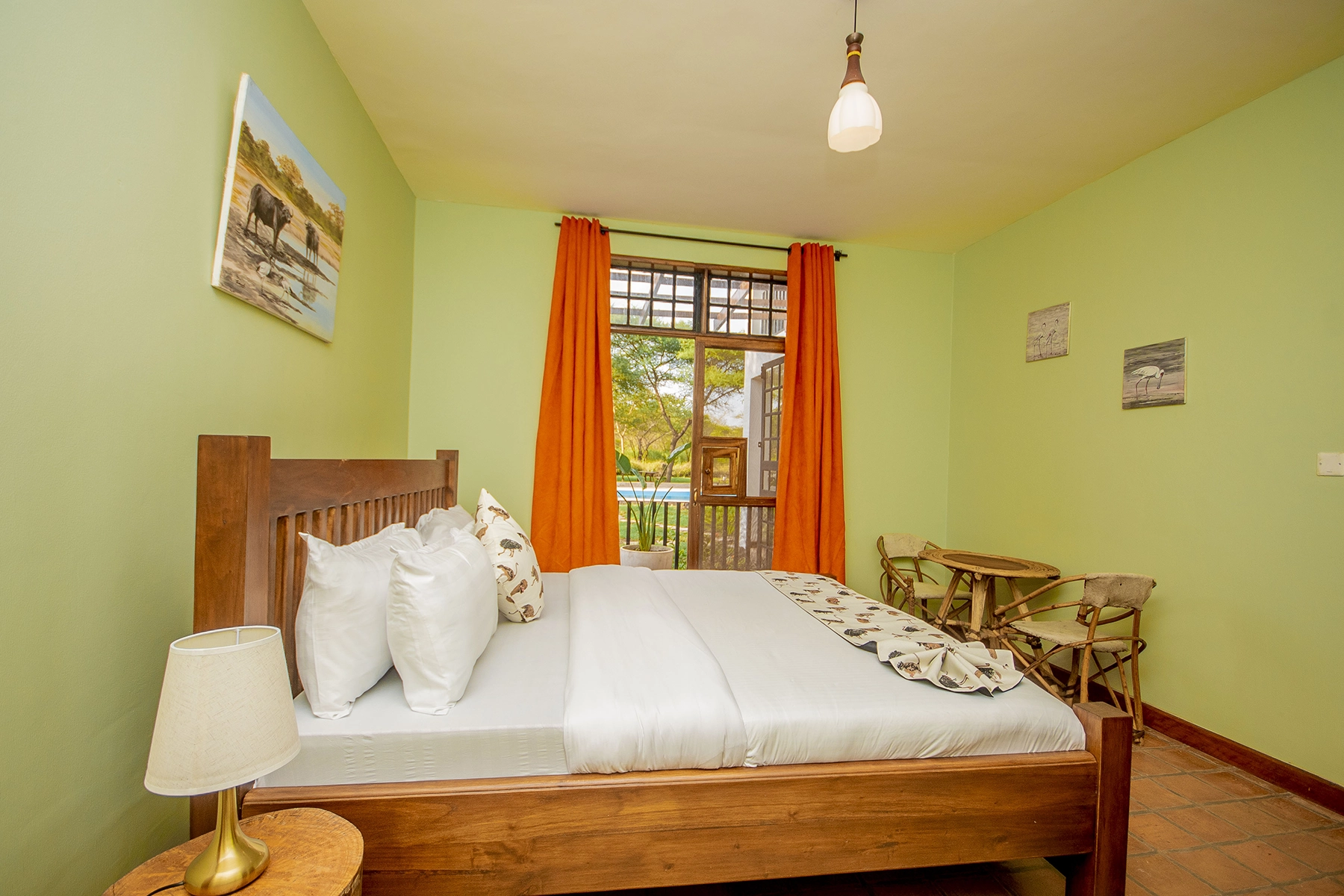 Bright double bedroom at Amani Farmhouse Tanzania with green walls, orange curtains, wooden double bed with bird-print cushions, and a balcony overlooking the pool and garden