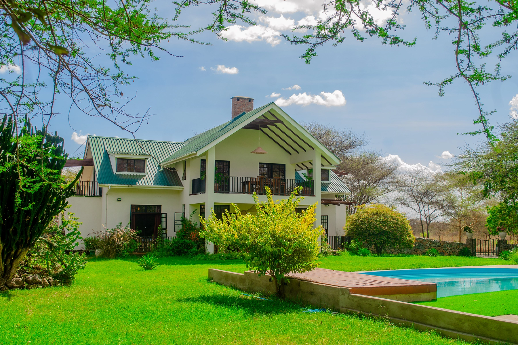 Two-storey white Amani Farmhouse with green roof, balcony, lush garden lawn, and outdoor swimming pool surrounded by acacia trees in Tanzania