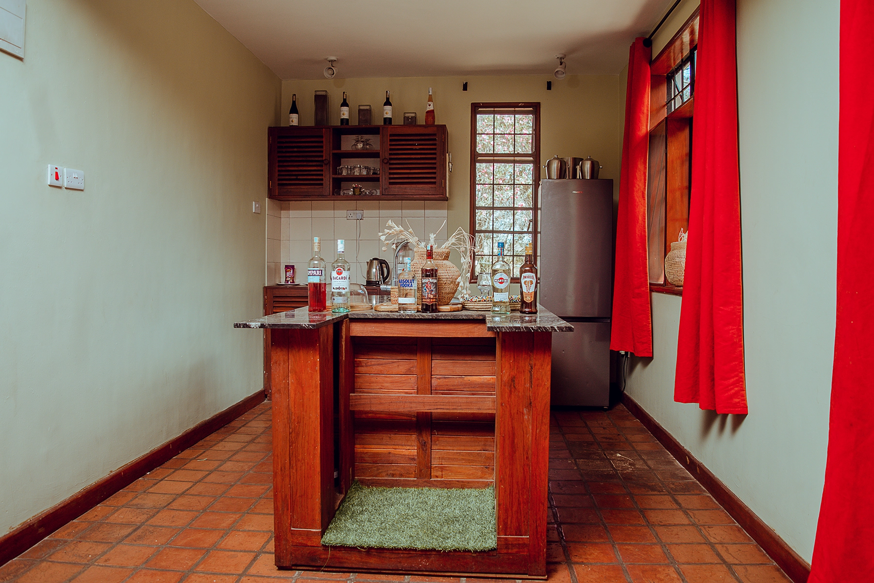 In-house bar area at Amani Farmhouse Tanzania with a marble-top wooden island, stocked spirits and wine bottles, wooden cabinetry, and a refrigerator