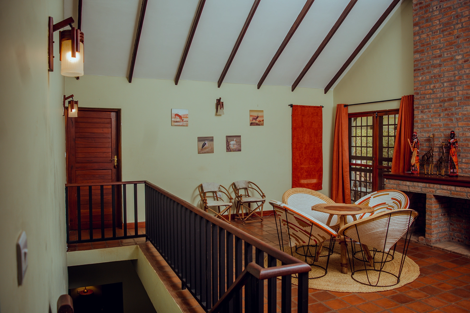 Upper-level lounge at Amani Farmhouse Tanzania viewed from the staircase, with woven basket chairs, African wildlife artwork, brick fireplace, and exposed timber beam ceiling