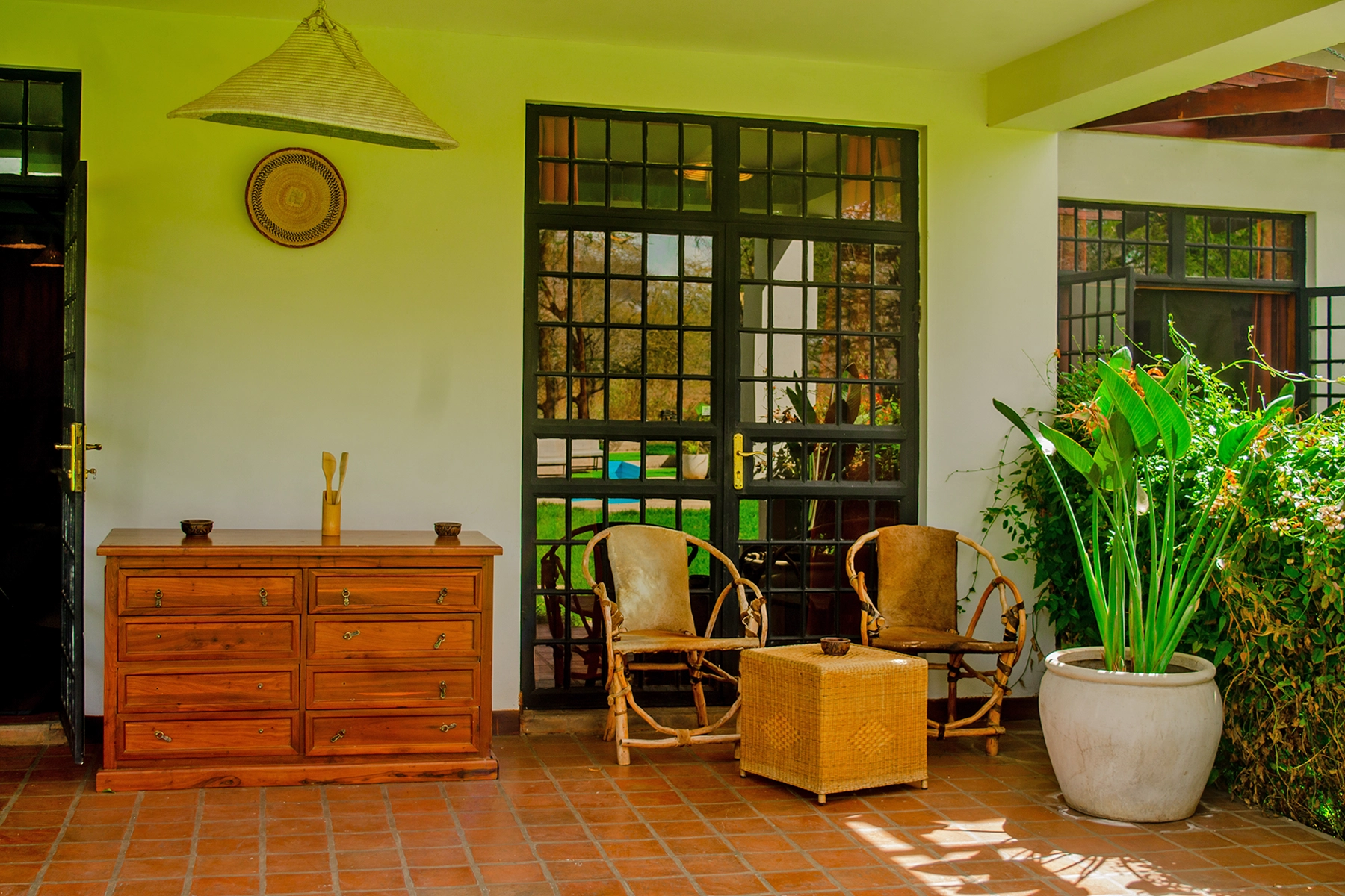 Shaded covered veranda at Amani Farmhouse Tanzania with terracotta tile floor, antique wooden dresser, rattan chairs, woven pendant light, and tropical garden plants