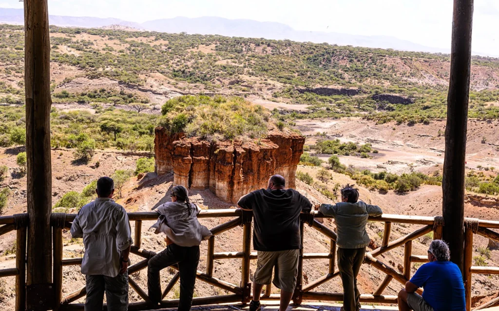 Visitors admiring the view of Olduvai Gorge from a wooden viewing platform, overlooking the eroded red rock formations and savanna landscape of the Cradle of Mankind in Tanzania