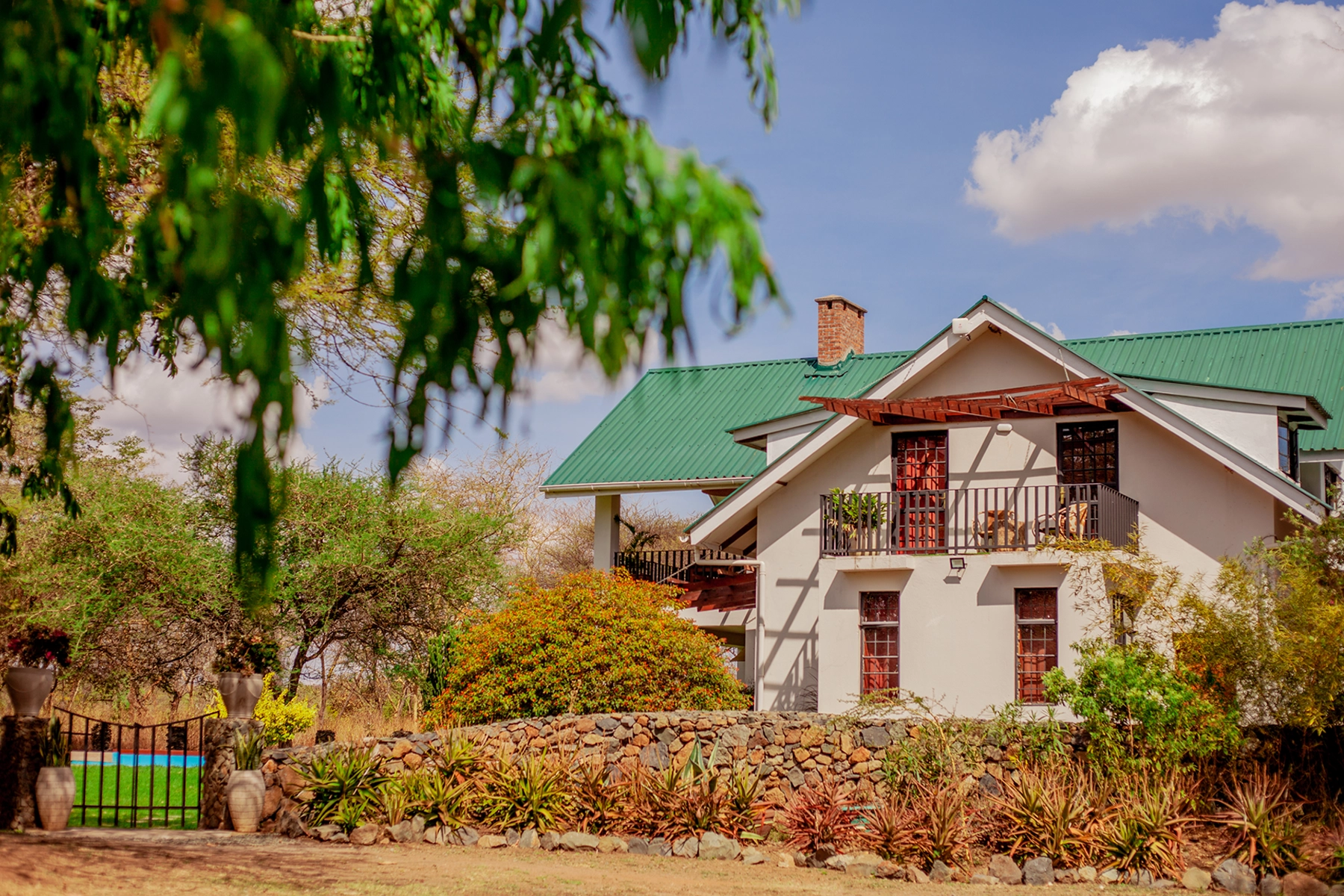 Exterior view of Amani Farmhouse in Arusha Tanzania, a white two-storey country house with a green roof, brick chimney, stone wall boundary, and lush garden framed by acacia trees with a glimpse of the swimming pool